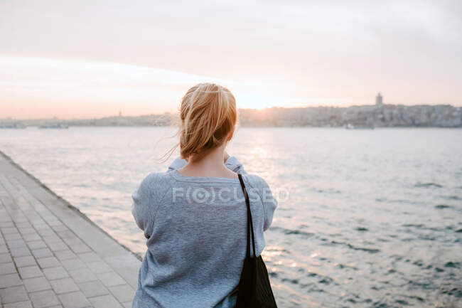 Back view of anonymous female traveler in casual outfit standing on embankment and admiring view of colorful amazing sunset — Stock Photo