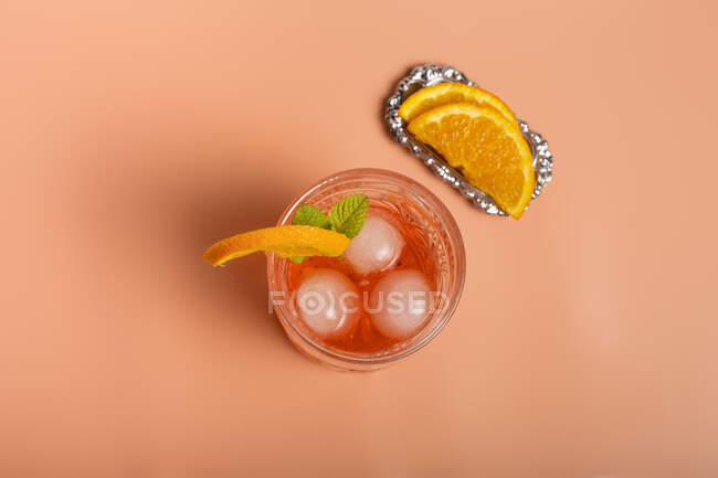 Вид зверху на алкогольний коктейль з кубиками льоду і гілочкою м'яти в склі, розміщеному на рожевому фоні з апельсиновими скибочками — стокове фото