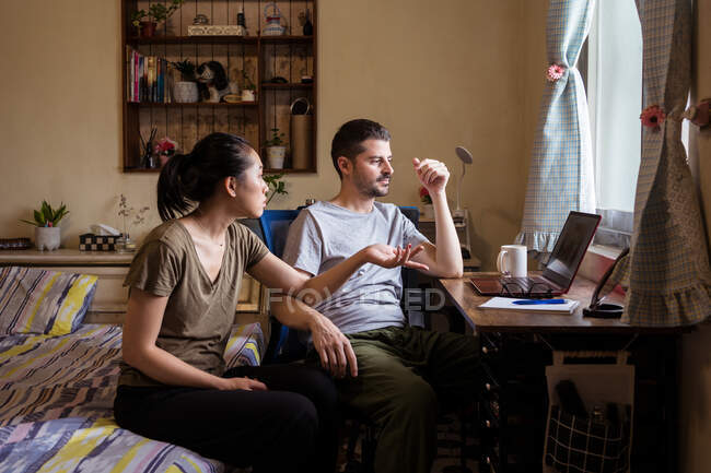Jeune couple en vêtements décontractés assis dans la chambre à coucher à table et en utilisant l'ordinateur ensemble tout en discutant vidéo — Photo de stock