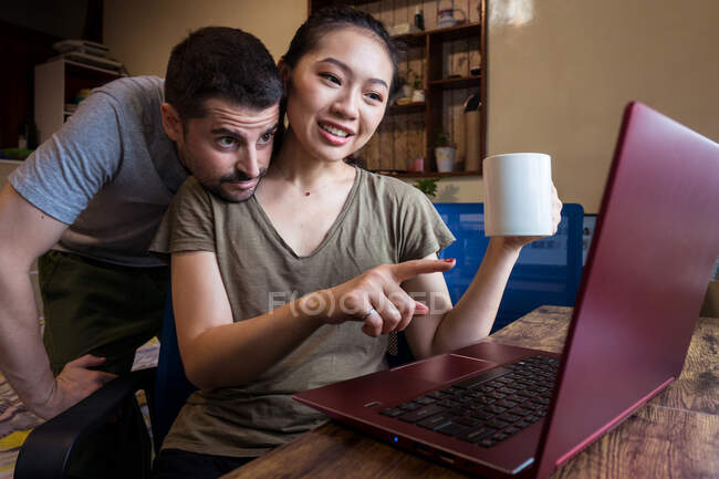 Bajo ángulo de novio feliz ayudando a novia asiática trabajando en el ordenador en casa mientras toma una bebida caliente en una taza - foto de stock