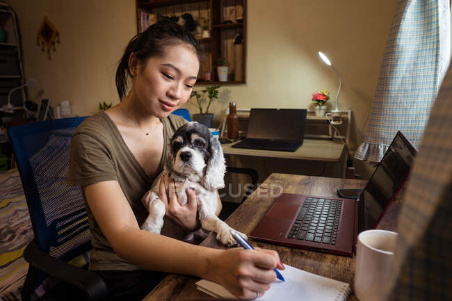 Вид сбоку сосредоточенной женщины-фрилансера, сидящей на стуле и делающей заметки на блокноте, держа в руках собаку, работающую дистанционно от дома — стоковое фото