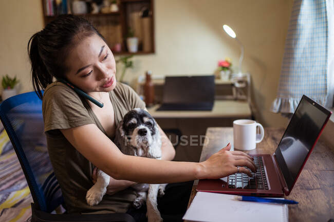 Freelancer femenina seria tomando notas en el ordenador portátil y hablando en el teléfono inteligente sobre el proyecto mientras está sentado en la silla con el perro y trabajando de forma remota - foto de stock