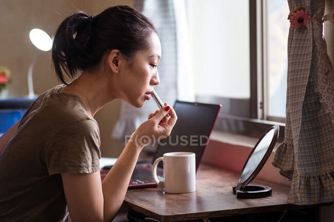 Vista lateral de una joven mujer étnica seria con ropa casual sentada en la mesa frente al espejo y aplicando lápiz labial - foto de stock