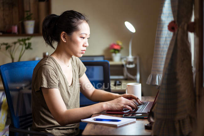 Vista lateral do freelancer feminino asiático em camiseta casual e óculos sentados à mesa e computador de navegação enquanto trabalhava no projeto online em casa — Fotografia de Stock