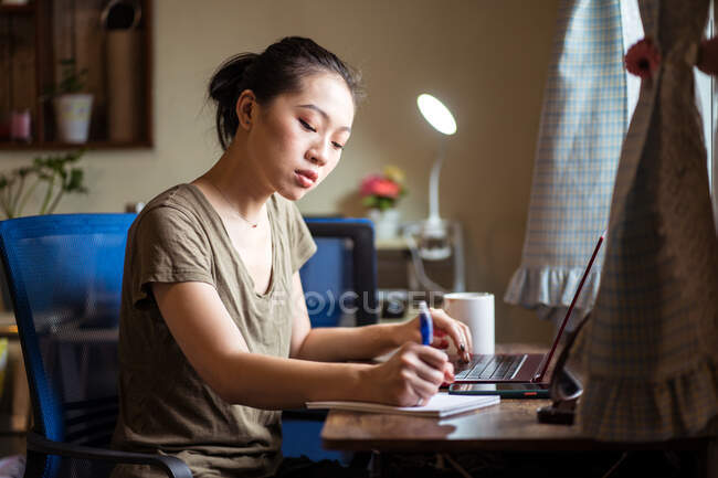 Libera professionista femminile concentrata in abbigliamento casual seduta a tavola con laptop e scrittura in blocco note mentre lavora sulla strategia aziendale in home office — Foto stock