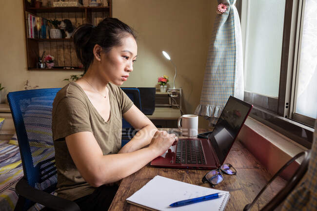 Vue latérale de la pigiste asiatique en t-shirt décontracté et lunettes assis à la table et l'ordinateur de navigation tout en travaillant sur le projet en ligne à la maison — Photo de stock