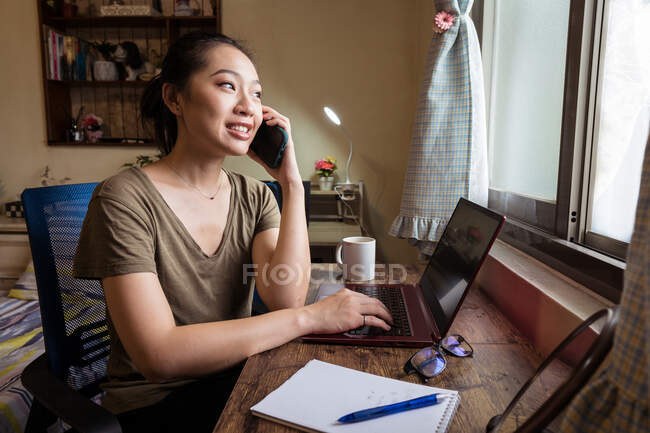 Side view of Asian female freelancer in casual t shirt sitting at table and browsing computer working on project online at home while speaking o the smartphone — Stock Photo