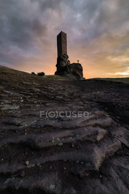 Vista de baixo ângulo do antigo monumento de pedra localizado na colina rochosa com silhueta de viajante anônimo em pé na rocha contra o céu nublado durante o pôr do sol — Fotografia de Stock