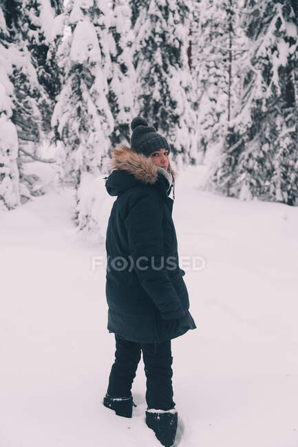 Вид сбоку на неузнаваемую женщину-путешественницу в теплой верхней одежде, стоящую на снежной тропе среди заснеженных елок в зимний день в Финляндии — стоковое фото