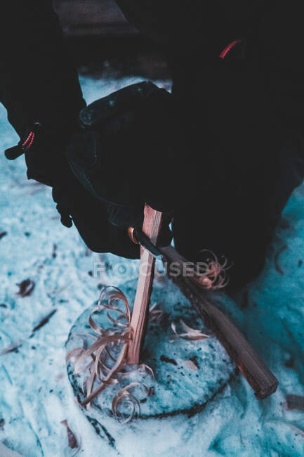 Сверху анонимный человек в теплой одежде режет щепки древесины острым инструментом на снегу в зимнем лесу в Финляндии — стоковое фото