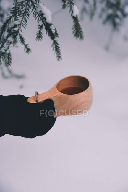 Боковой вид обрезанной неузнаваемой руки в теплой перчатке с чашкой горячего напитка, стоящей в снежном лесу в зимний день в сельской местности Финляндии — стоковое фото