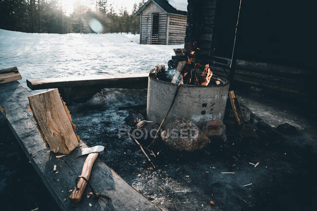 Hoguera ardiente y tronco con hacha colocada cerca de una pequeña cabaña de leñador en el bosque nevado en el día de invierno en el campo de Finlandia - foto de stock