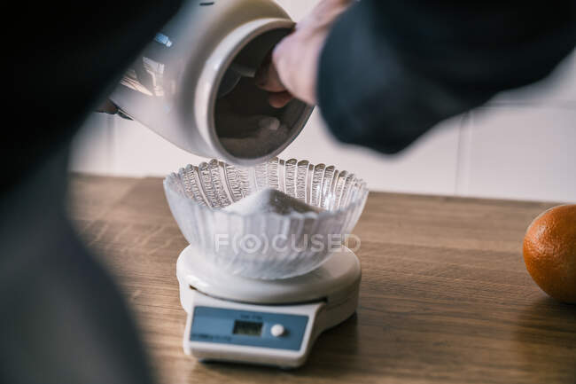 Zugeschnittene, unkenntlich gemachte weibliche Hand, die Zuckerdose verschüttet und Zucker mit elektronischer Küchenwaage mit Glasschale auf Holztisch beschwert — Stockfoto