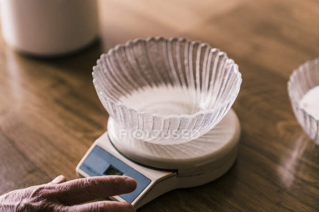 Cropped unrecognizable female hand weighting with electronic kitchen scale with glass bowl on top on wooden table — Stock Photo