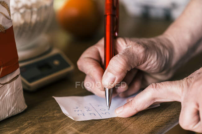 З понад врожаю анонімна зріла жінка в фартусі робить розрахунок на папері, готуючи інгредієнти для тіста на домашній кухні. — стокове фото