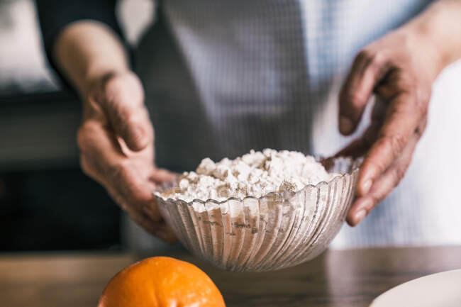 Unerkennbarer Konditor in Schürze steht in der Küche und hält Glasschale mit Mehl, während er Teig in der Nähe von Bir Orange zubereitet — Stockfoto