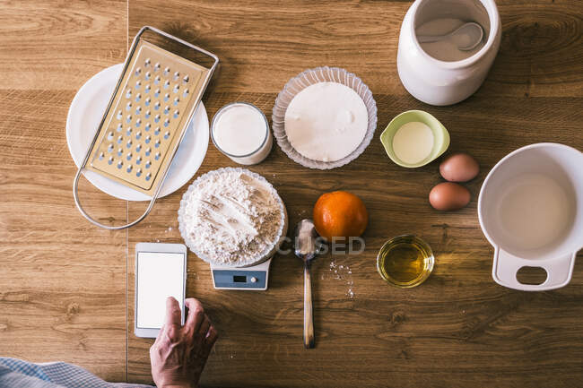 Vista dall'alto della ricetta anonima di navigazione femminile su smartphone e pesatura della farina di frumento su bilance elettroniche mentre si preparano ingredienti per la pasticceria fatta in casa in cucina — Foto stock