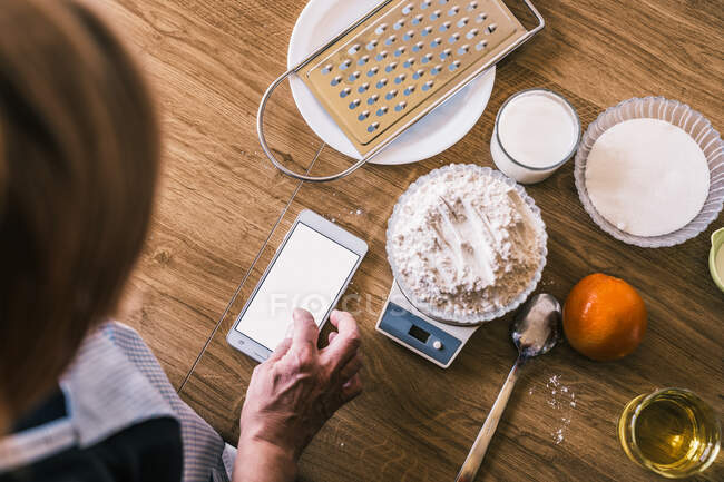 Vue du dessus de la recette de navigation féminine anonyme sur smartphone et pesant la farine de blé sur des balances électroniques tout en préparant des ingrédients pour la pâtisserie maison dans la cuisine — Photo de stock