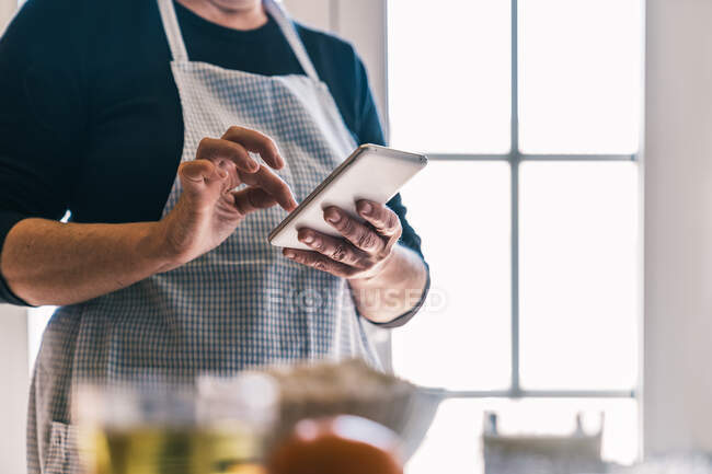 Вид сбоку на обрезную самку в фартуке, просматривающую смартфон и ищущую еду, стоя на светлой домашней кухне — стоковое фото