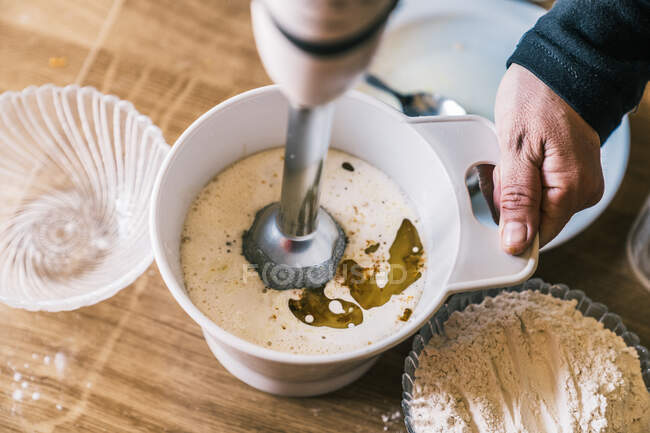 Dall'alto raccolto casalinga anonima utilizzando mixer durante la preparazione di pasta per muffin dolci al bancone della cucina con ingredienti per la ricetta — Foto stock