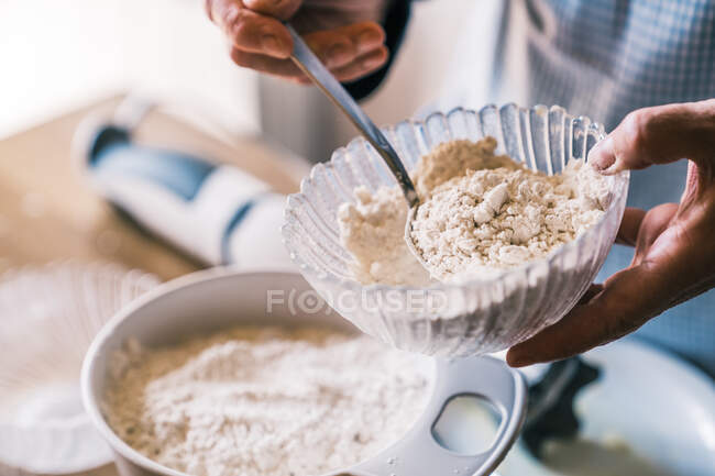 From above unrecognizable confectioner in apron standing in kitchen with glass bowl and adding flour with spoon while preparing dough for muffins — Stock Photo