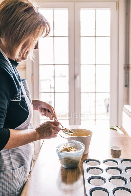 Вид сбоку на пожилую домохозяйку, стоящую за столом с подносом для кексов и наливающую тесто ложкой в кексовые подкладки — стоковое фото