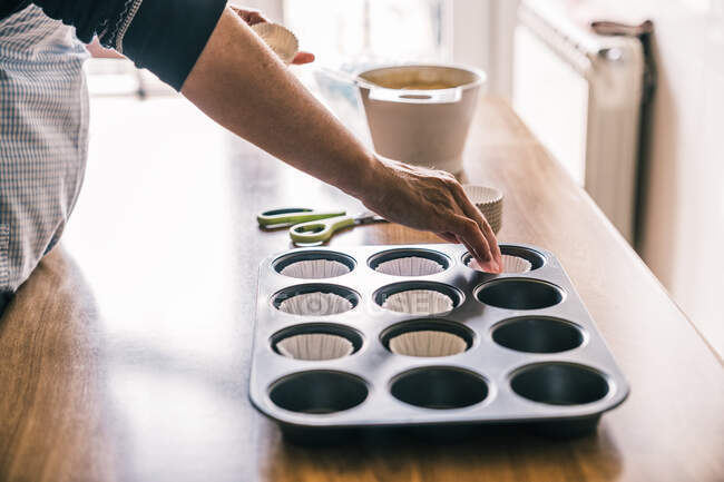 Seitenansicht eines betagten Konditors in Schürze, der am Tisch mit Muffin-Tablett und Papiereinlagen steht, während er Geschirr zum Kochen zubereitet — Stockfoto