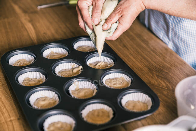 De cima colheita confeiteiro feminino de pé à mesa com bandeja de muffin e enchimento de forros de papel com massa na cozinha em casa — Fotografia de Stock