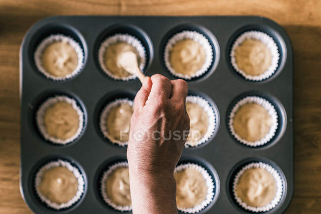Vista superior da dona de casa idosa anônima em pé à mesa com bandeja de muffin e derramando massa com colher em forros de cupcake — Fotografia de Stock