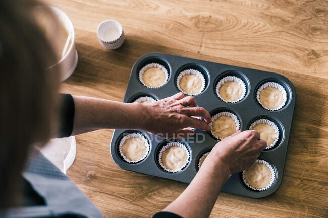 Crop pasticceria femminile in piedi a tavola con vassoio di muffin fissaggio rivestimenti di carta con pastella in cucina a casa — Foto stock