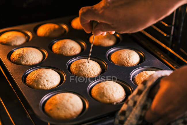 Crop confectioner checking dough in muffins with metal stick while cooking delicious homemade dessert in kitchen — Stock Photo
