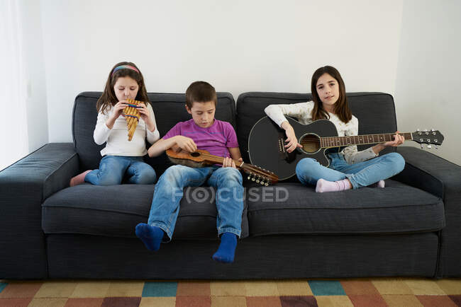 Niño y niñas positivos con ropa casual sentados en un cómodo sofá juntos y tocando instrumentos de música mientras pasan tiempo juntos en casa - foto de stock