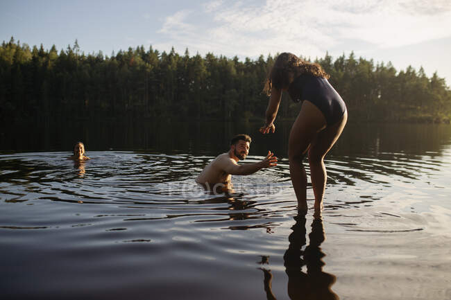 Вид ззаду анонімної молодої стрункої леді в купальнику, що йде у воду спокійного лісового озера, щоб плавати друзям — стокове фото