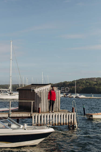 Male tourist in red warm jacket standing alone on wooden pier with boats and yachts on sunny day in weekend and looking away — Stock Photo