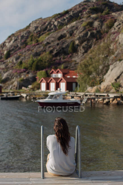Обратный вид анонимной туристки в повседневной теплой одежде, сидящей на деревянной набережной и любующейся спокойным видом на лодку и дома — стоковое фото