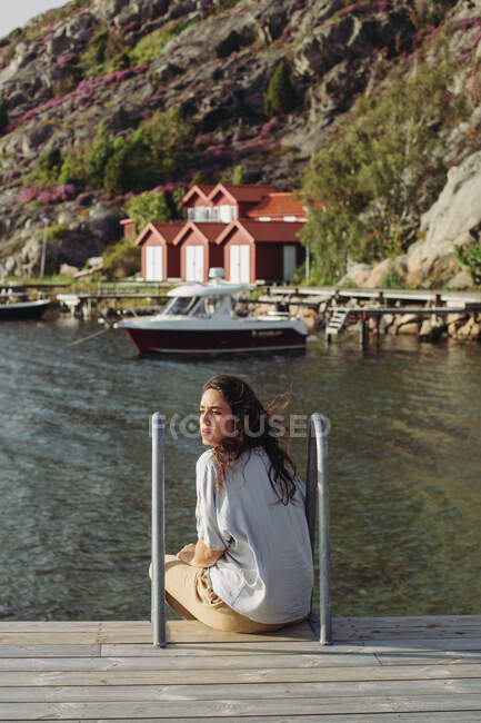 Vue arrière du touriste féminin en vêtements décontractés assis sur une promenade en bois regardant loin admirant une vue calme sur le bateau et les maisons — Photo de stock