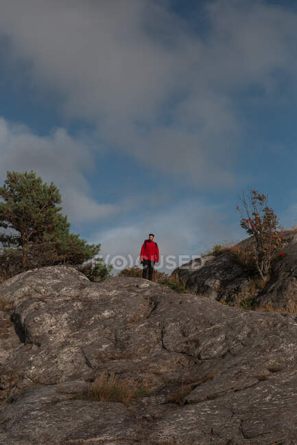 Turista masculino em casaco vermelho e com mochila andando na encosta rochosa segurando câmera fotográfica e tirando fotos da bela paisagem no dia nublado — Fotografia de Stock