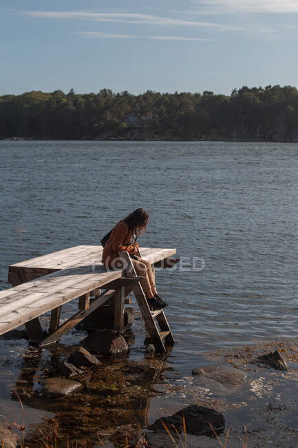 Turista femenina en ropa de abrigo y con mochila descansando en muelle de madera cerca de un lago tranquilo y mirando al agua en un día soleado - foto de stock