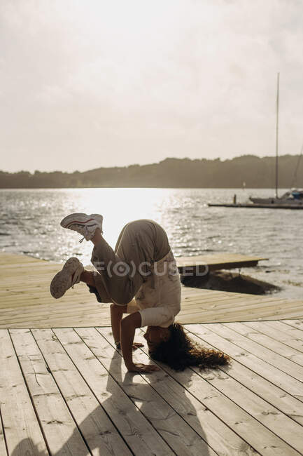 Vista lateral de la bailarina en ropa casual y zapatillas blancas de pie en la cabeza en el paseo marítimo de madera en el día soleado durante las vacaciones - foto de stock