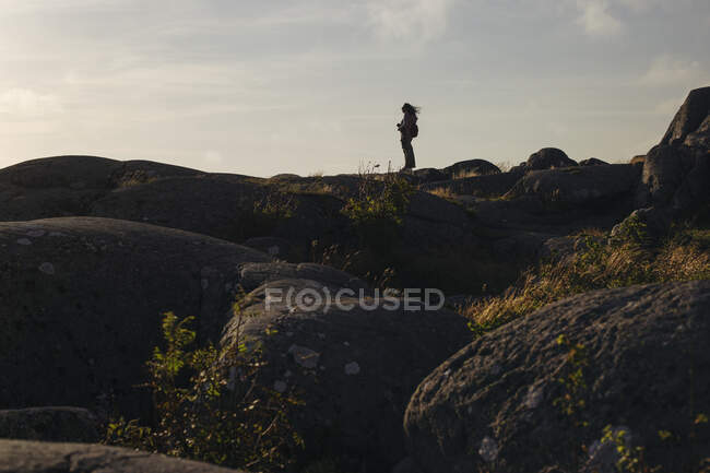 Vue latérale silhouette de touriste femelle avec sac à dos debout sur la colline rocheuse avec appareil photo admirant beau paysage par temps nuageux — Photo de stock