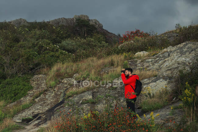 Vista lateral del turista masculino en chaqueta roja y con la mochila de pie en la ladera rocosa y tomar fotos del paisaje en el día nublado - foto de stock