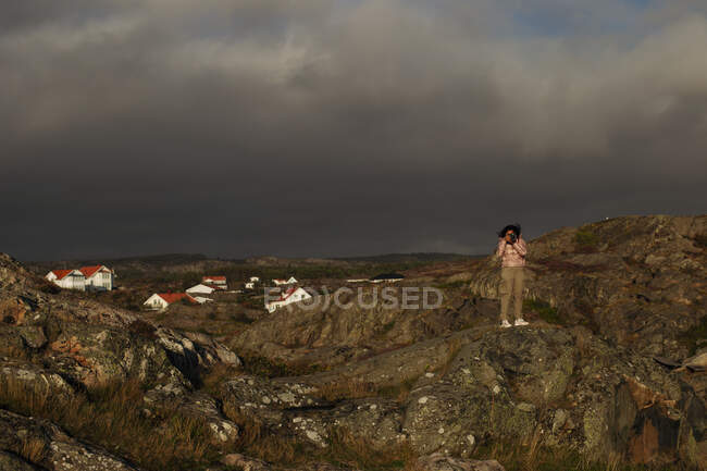 Anonimo viaggiatore femminile in tuta casual in piedi sulla riva rocciosa e scattare foto con macchina fotografica nella giornata ventosa e nuvolosa — Foto stock