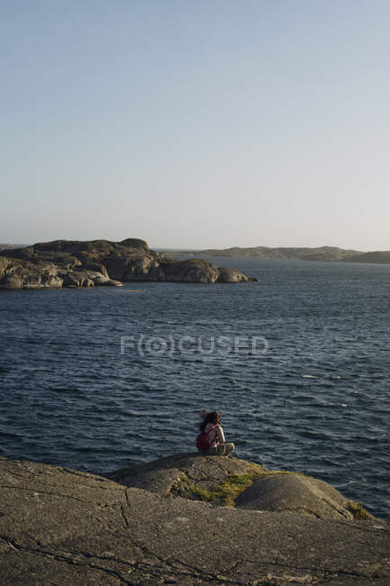 Back view of anonymous female tourist in casual warm outerwear and with backpack sitting on edge of rocky seashore and admiring amazing view of sea on windy day — Stock Photo