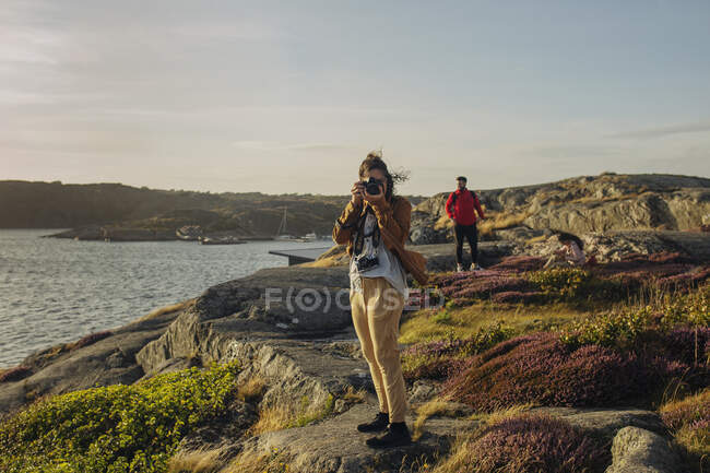 Turista femenina anónima en ropa casual de pie sobre piedra y tomando fotos en cámara fotográfica mientras camina en la costa rocosa con su amigo - foto de stock