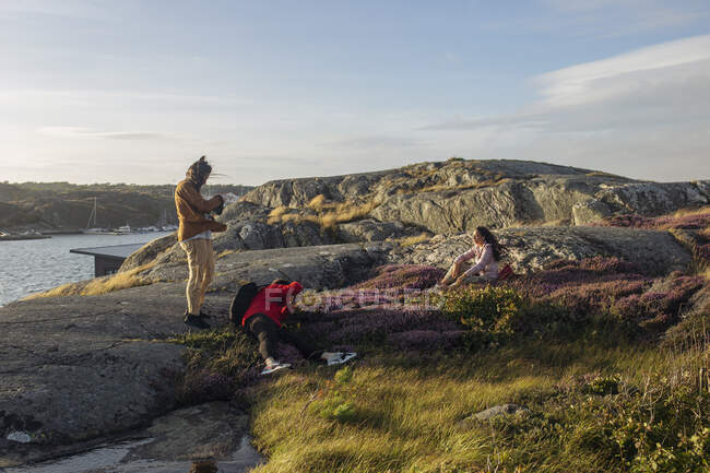 Group of tourist in casual clothes standing on stone and taking pictures of friend on photo camera on rocky seashore — Stock Photo