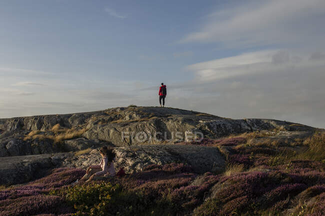 Back view of anonymous couple of travelers walking on rocky cliff covered with grass and flowers and admiring picturesque landscape — Stock Photo