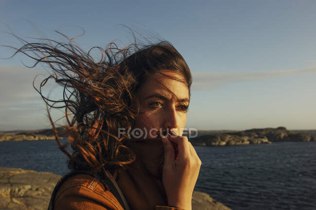 Beautiful young female traveler in casual clothes standing on rocky seashore with hair blowing in wind and looking away dreamily — Stock Photo