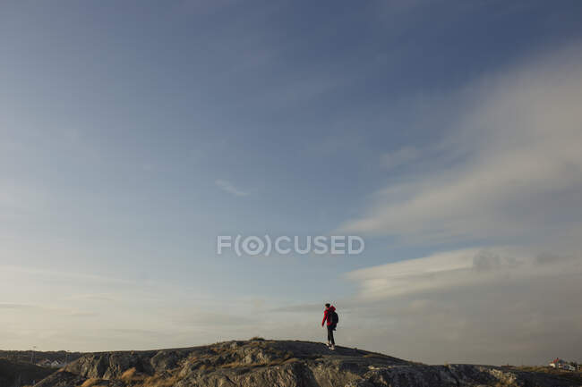 Vista lateral del turista masculino en chaqueta roja y con mochila caminando sobre una colina rocosa sosteniendo una cámara fotográfica y tomando fotos de un hermoso paisaje en un día nublado - foto de stock