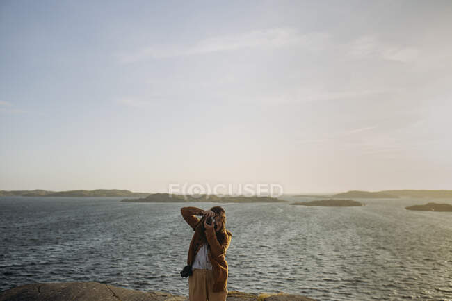 Анонімні жінки-туристки в повсякденному одязі, що стоять на скелястій скелі на березі моря і фотографуються, насолоджуючись дивовижним морським пейзажем під час відпустки — стокове фото