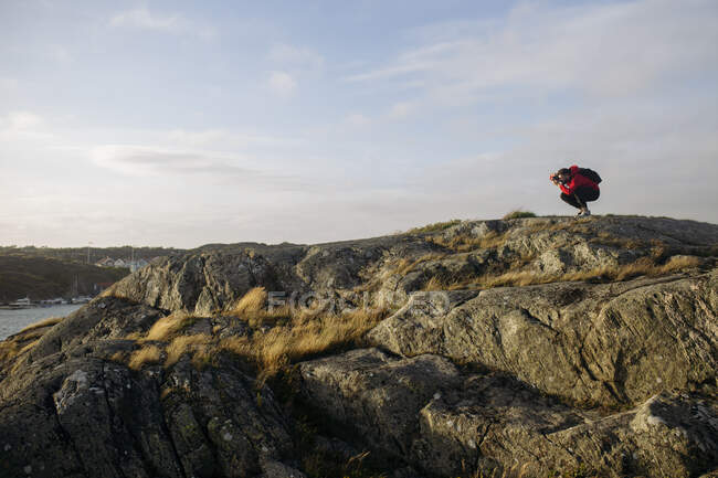 Vista lateral del viajero masculino con mochila en ropa casual agachándose mientras toma una foto del paisaje del lago con cámara fotográfica en la superficie rocosa - foto de stock